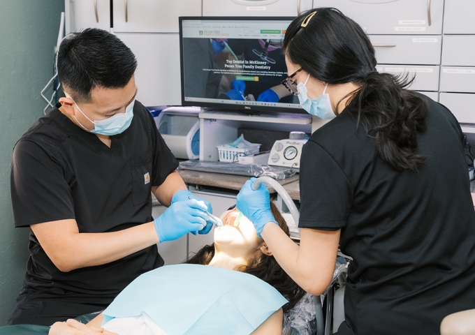 Dr. Peter J. Kim and a masked dental assistant at Pecan Tree Family Dentistry in McKinney, TX, perform a same-day dental procedure on a reclining patient, with digital X-rays displayed on a monitor behind them.